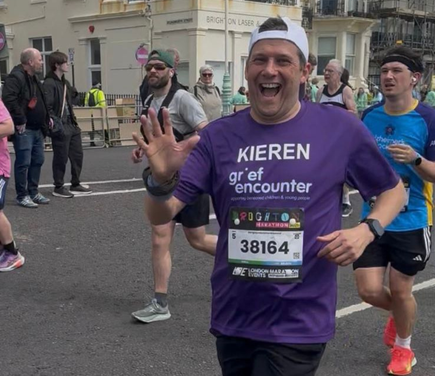 Runner in a purple shirt joyfully waves during a race, with spectators and other runners in the background. The atmosphere is energetic and lively.