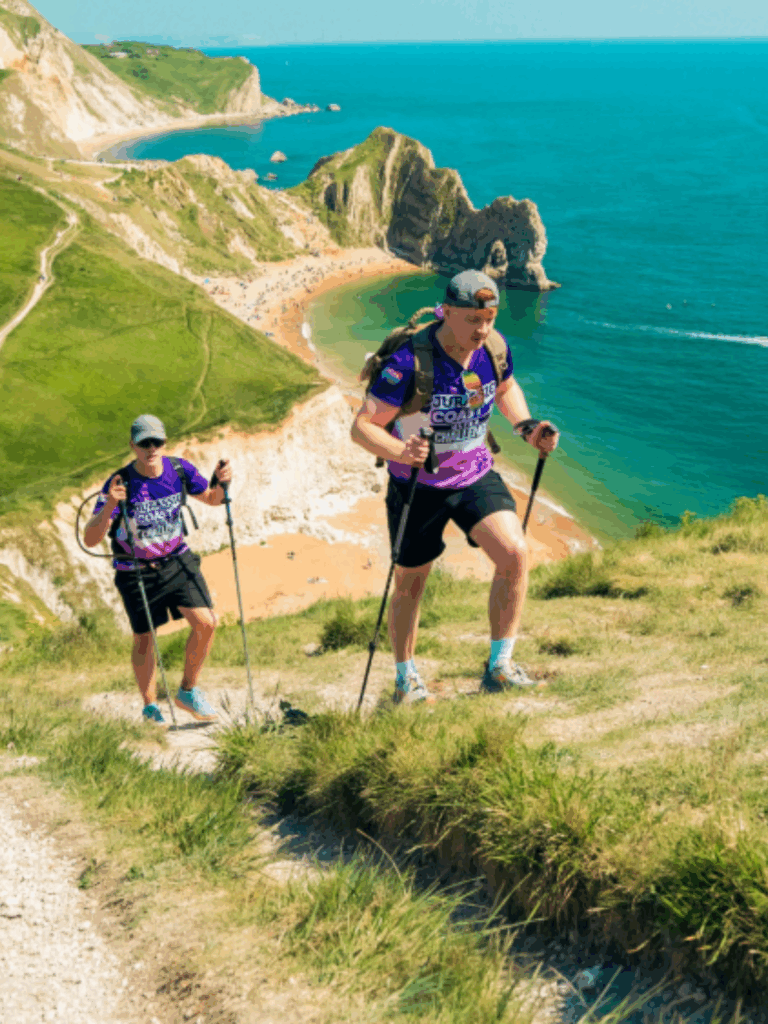 Two people in athletic gear hike uphill with trekking poles along a coastal path overlooking a beach and cliffs on a sunny day.