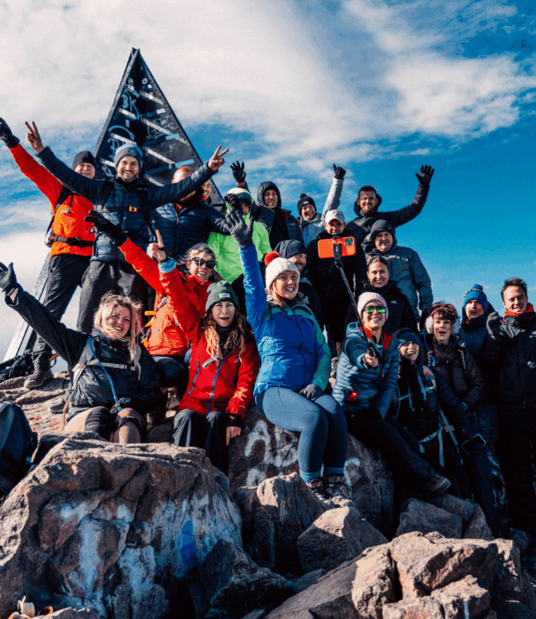 A group of climbers, dressed in colourful winter gear, celebrate atop a rocky peak under a bright, partly cloudy sky, with a triangular summit marker.