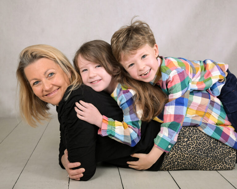 An older white women in a black top is lying down with two children on her back. A young white girl with long brown hair and a young white boy with short brown hair. Both children are wearing multi coloured shirts.