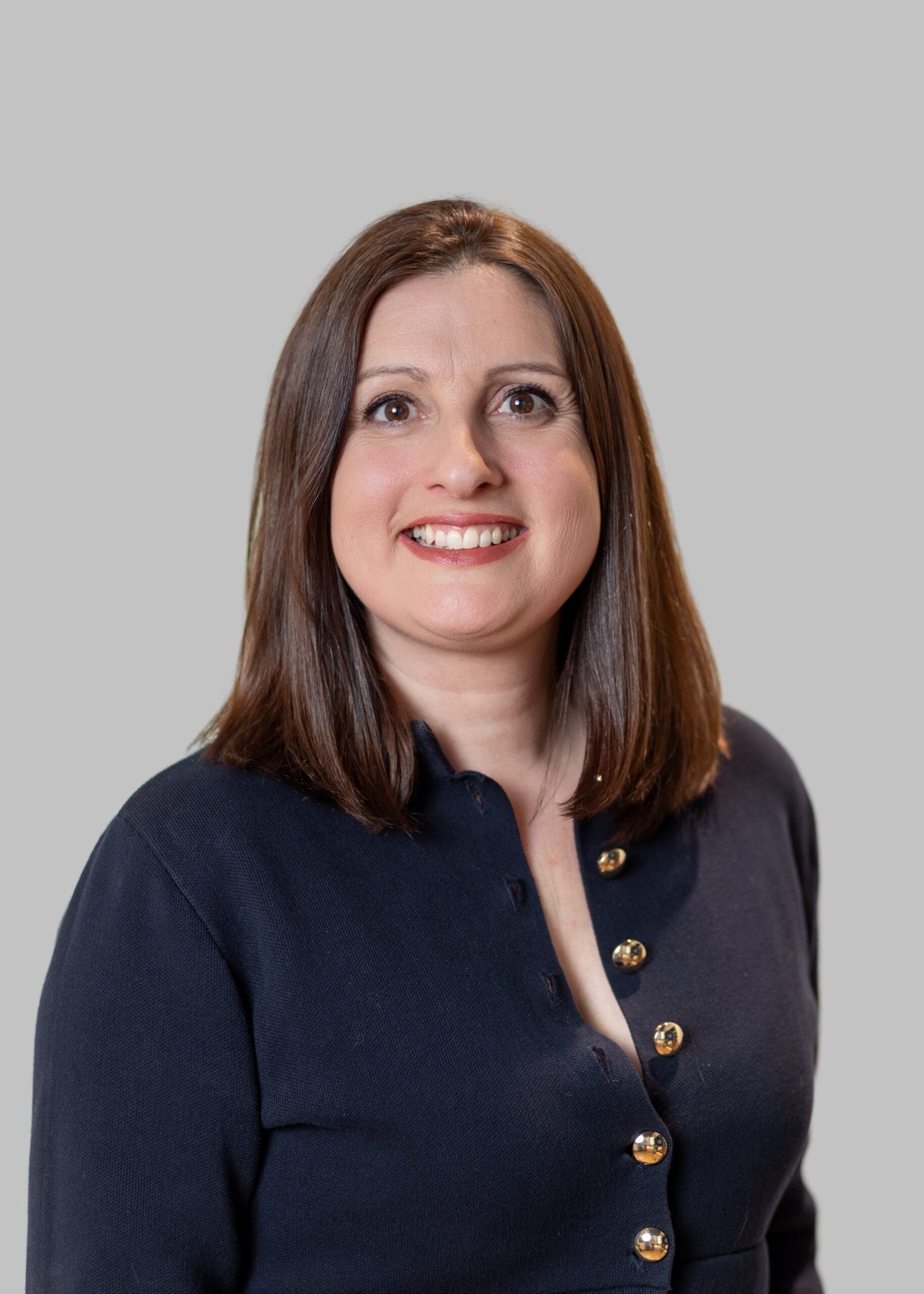 Woman with straight brown hair wearing a navy blue top with gold buttons, smiling, posed against a plain light gray background.