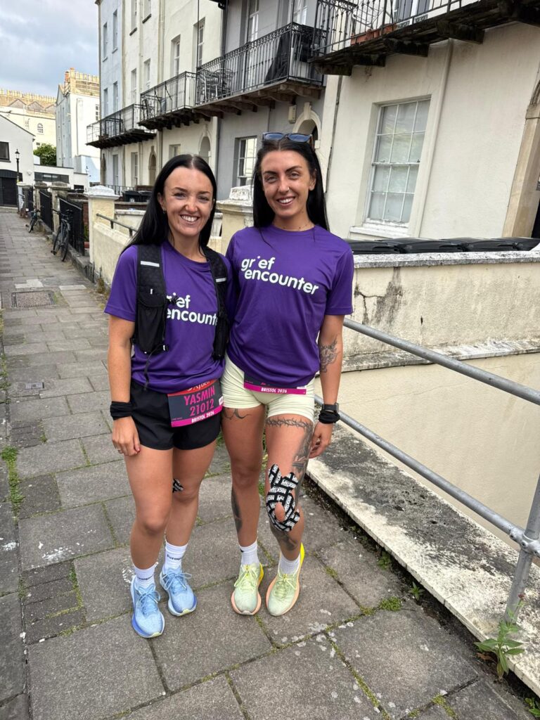 Two young white women, standing together wearing purple Grief Encounter running tops. They are preparing to take on the Bristol Half Marathon.